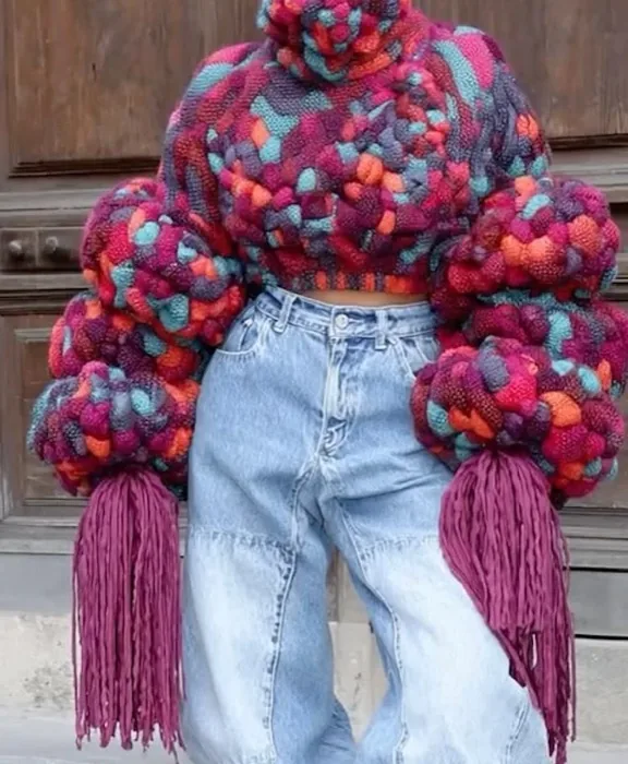 Maximalist chunky knit sweater with oversized multicolor pom-pom sleeves and fringe, representing Playscape's playful volume and texture